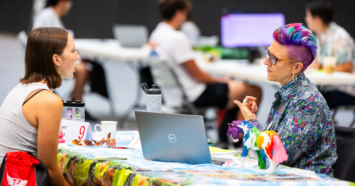 A student sits across from and chats with her advisor at a table that's decorated with colorful tablecloth.