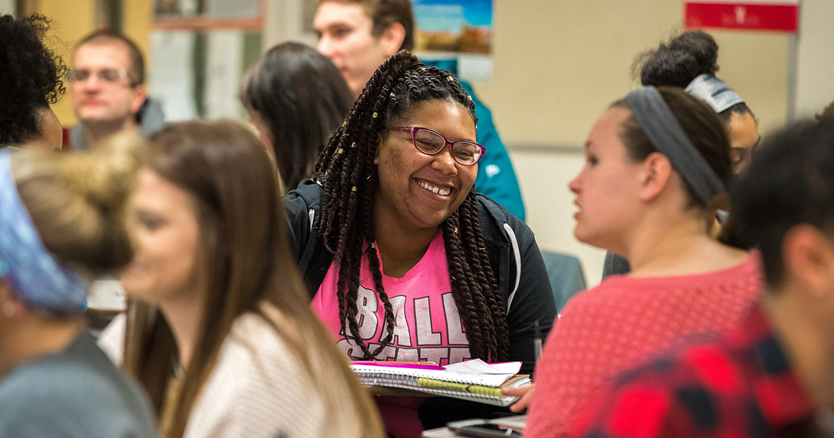 A girl surrounded by other students smiles in the middle of a classroom