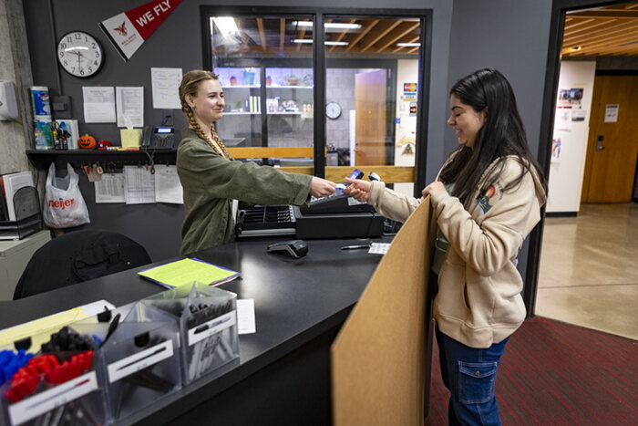 Two students shake hands across a desk while one holds a large brown sheet of paper