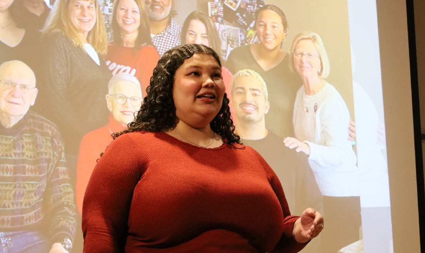 Female student with red shirt and dark hair in front of a projector screen giving a speech. A group photo is displayed in the background on the projector