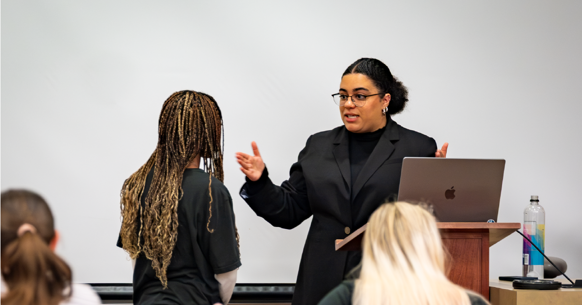 Faculty member speaking with a student at the front of a classroom while other students sit.