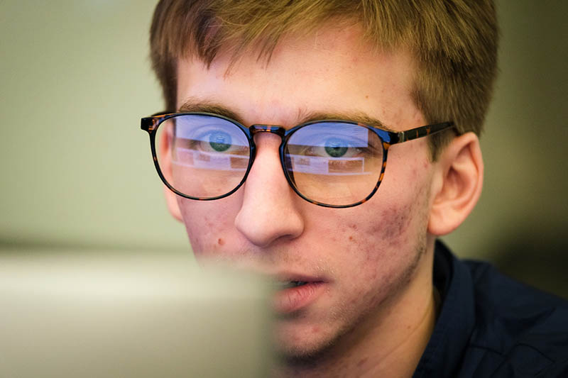 a student looks closely at a computer screen, the light reflecting off his glasses