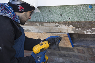 A student in a hard hat attaches a board to the side of a house with a yellow drill.