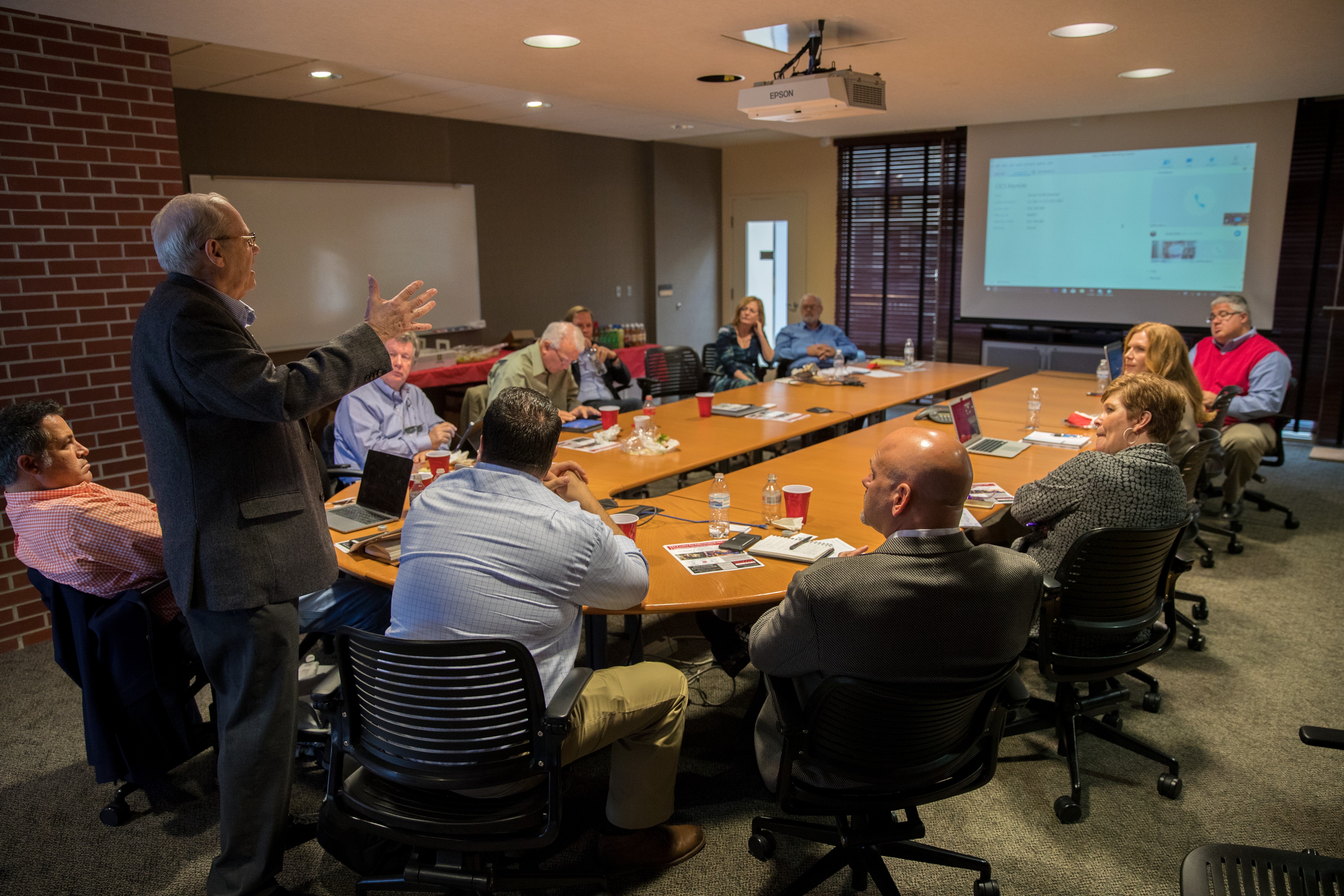 Large oval conference table with professionals seated around it. One gentlement at the end in a suit is standing and speaking and a presentation is displayed on the far end of the photo.
