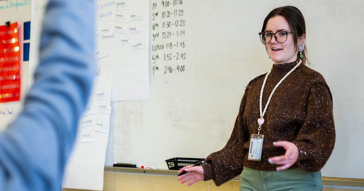 A young woman wearing glasses, a brown sweater, and a lanyard with an ID card stands in front of a whiteboard while teaching a class