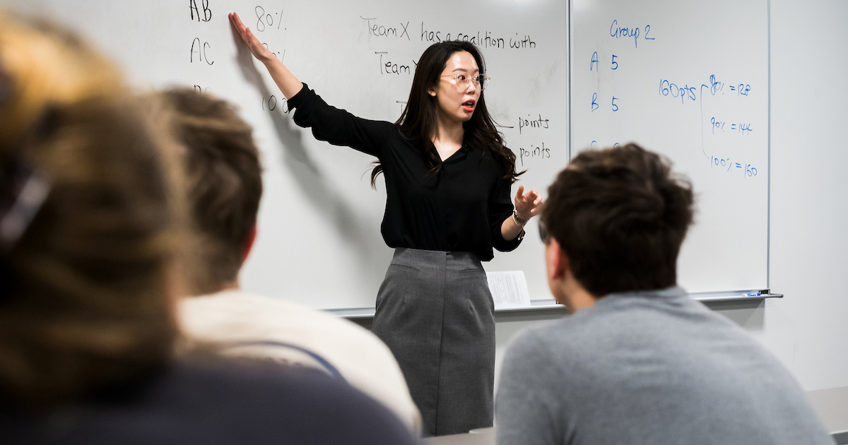 woman points at a whiteboard and speaks to a classroom of people