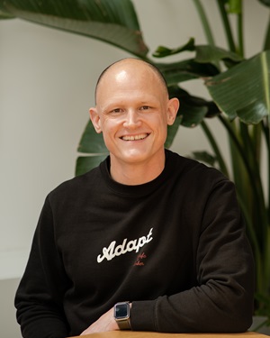 Smiling man with shaved head wearing a black sweatshirt Plant in background