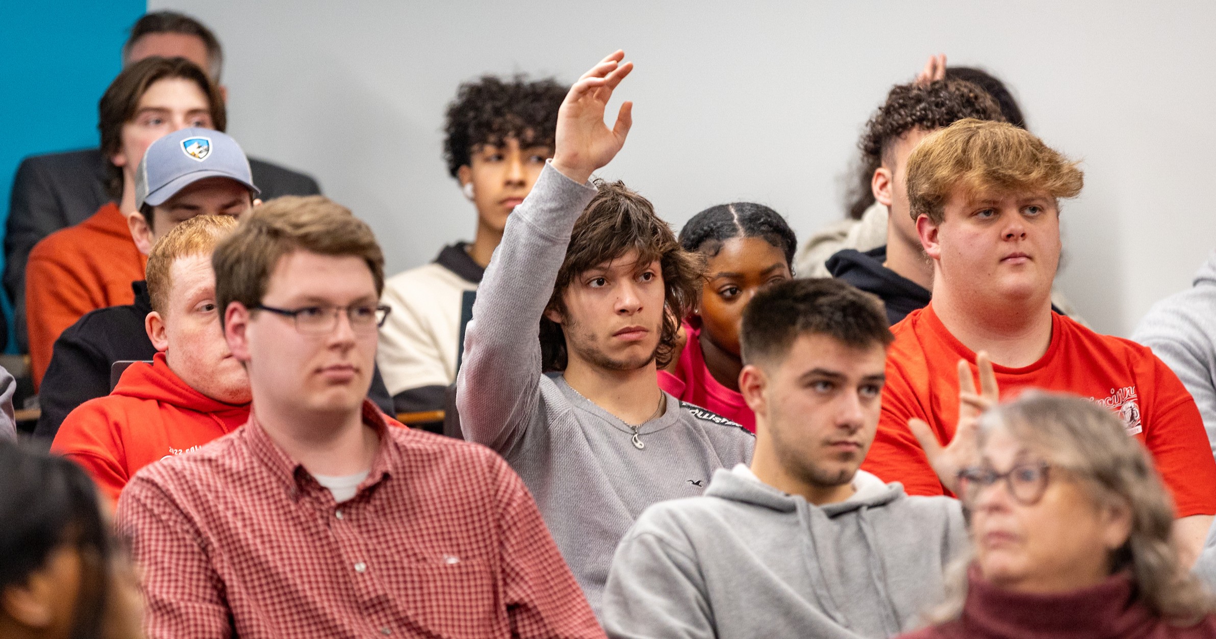 small group of students sitting in a classroom. One male student in the middle of the picture raising his hand