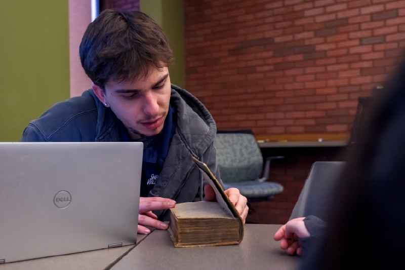 A student examines an old, worn book while seated at a table with a laptop, as classmates gather nearby in a collaborative learning space.