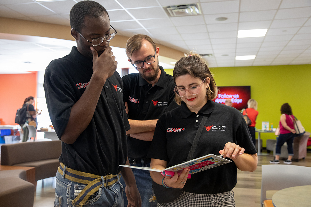 Three Ball State students in black polos read a book. 