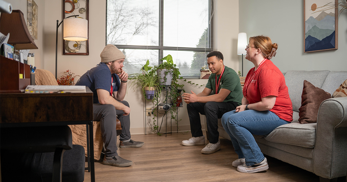 Three students sit conversing in an office. Two sit on a couch on the right across from one sitting on a chair on the left. Artwork and plants adorn the walls of the office. A window with open blinds is in the background. A desk with an open book is in the foreground. 