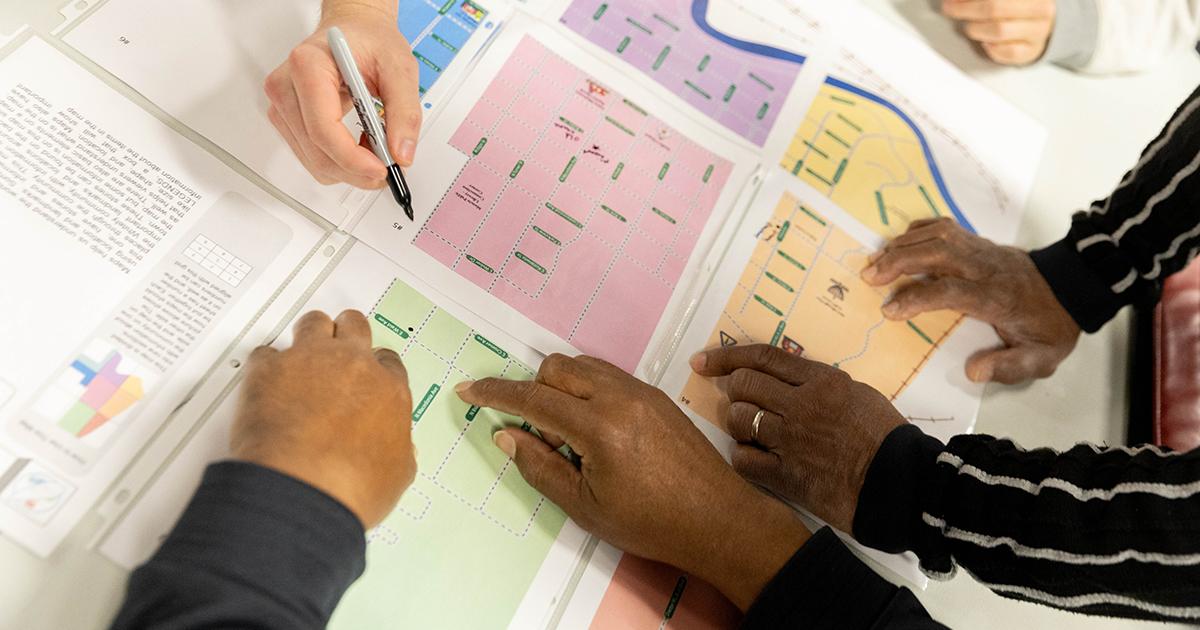 Several pairs of hands rest on a map of the Whitely Neighborhood in Muncie, Indiana. One hand holds a black sharpie. The map is broken into regions of different colors. Some of the hands point to a spot at N Macedonia Ave in a green region of the map for a Northwest area of the Whitely Neighborhood.