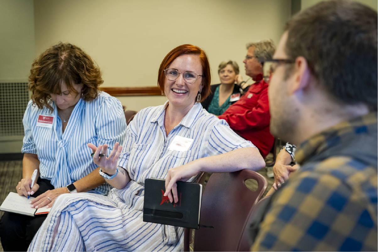 A smiling participant gestures while speaking during a small group discussion at a workshop or meeting, holding a notebook as another attendee takes notes nearby and others listen in the background.