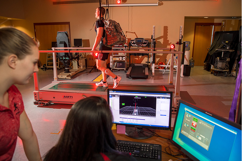 Two students look-on as a man runs on a treadmill.