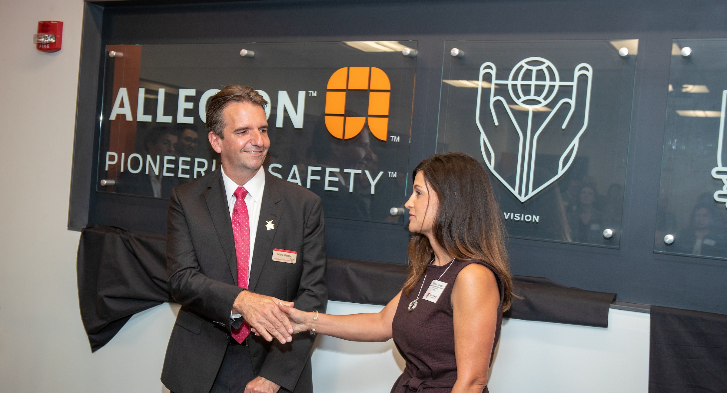 A man and a woman shake hands in an office, in front of a sign that reads, ALLEGION PIONEERING SAFETY.