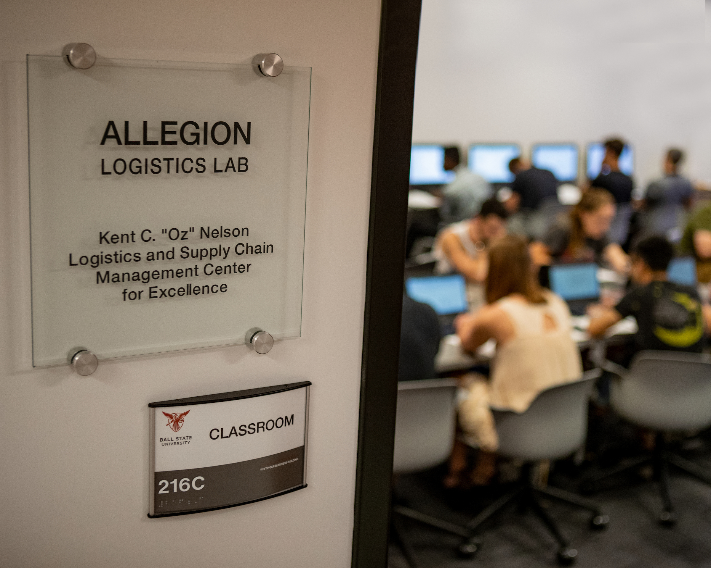 A sign outside room 216C identifies it as the Allegion Logistics Lab at Ball State University, with students visible working at computers inside the classroom.
