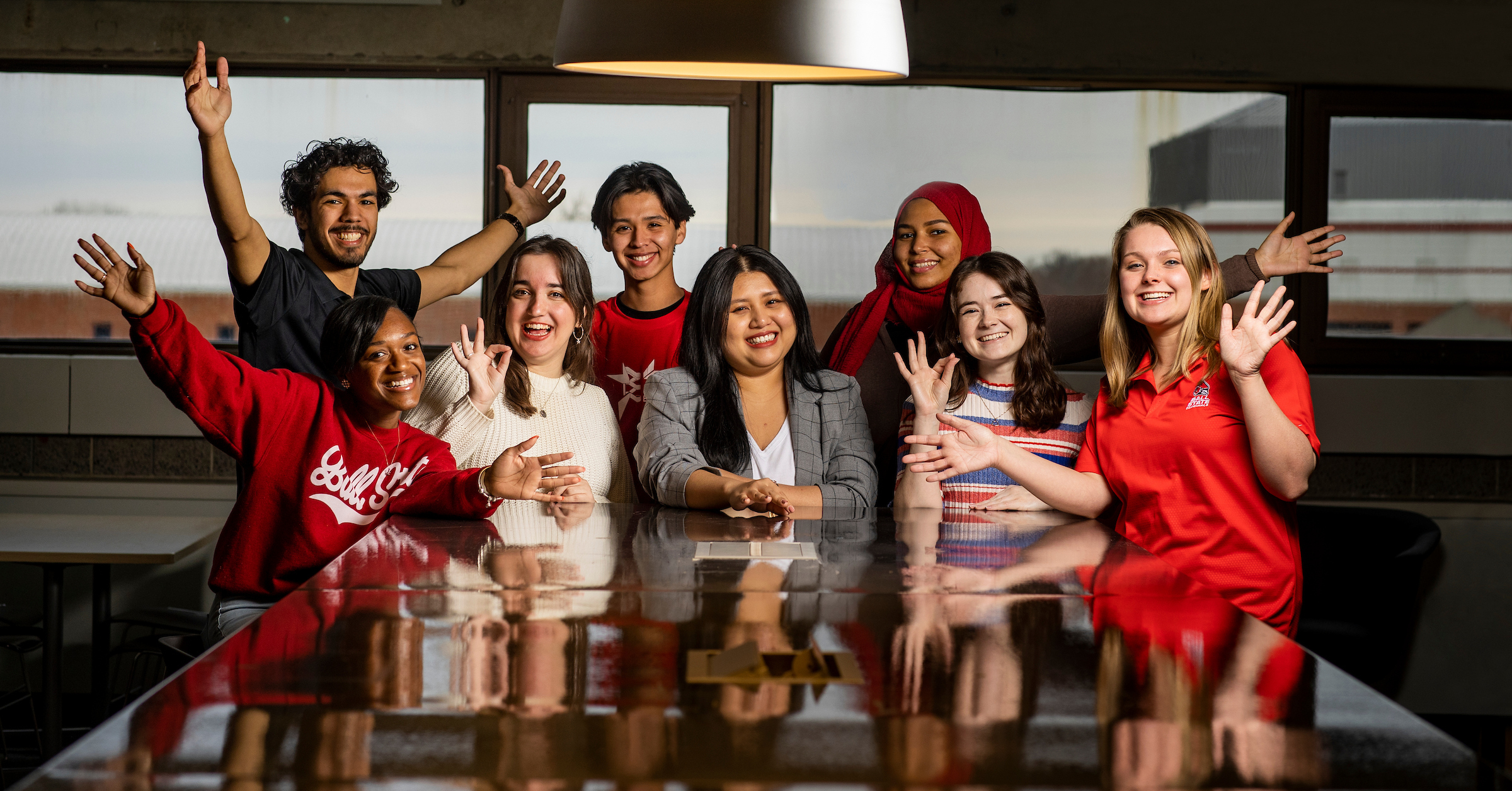 A group of eight smiling young adults sit and stand around a table indoors, waving and gesturing enthusiastically toward the camera. They appear diverse in ethnicity and style, wearing mostly red and casual clothing. Large windows and a hanging light fixture are visible behind them, and their reflections can be seen on the glossy tabletop in the foreground.