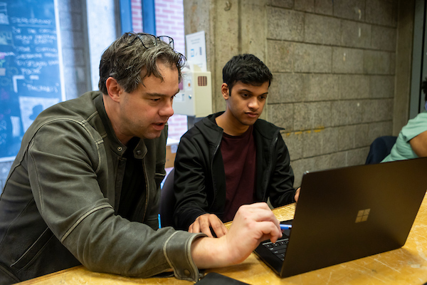  Gemini said Two men sit at a wooden table in an industrial-style room, focusing on a black laptop screen. One man, wearing a grey jacket, points at the screen while the other man, in a black hoodie, watches closely. A grey concrete block wall and a chalkboard with white writing are visible in the background.