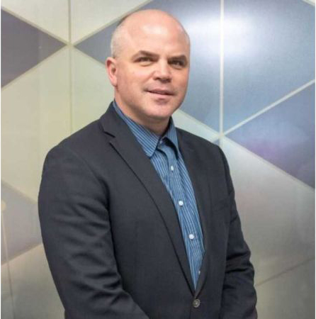 A professional headshot of Aaron Jones with a shaved head and a light complexion. He is smiling slightly and wearing a dark blazer over a blue and white pinstriped button-down shirt. The background features a geometric pattern of light blue and grey triangles.