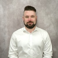 A headshot of Ian Francis with dark hair styled in a fade with a quiff on top and a well-groomed beard and mustache. He is wearing a crisp white button-down shirt and looking directly at the camera with a neutral expression. The background is a textured light grey wall.