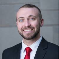 A professional headshot of Jason Fry smiling with short brown hair and a groomed beard. He is wearing a dark suit jacket, a white dress shirt, and a vibrant red tie. The background is a blurred, neutral-toned stone or tile wall.