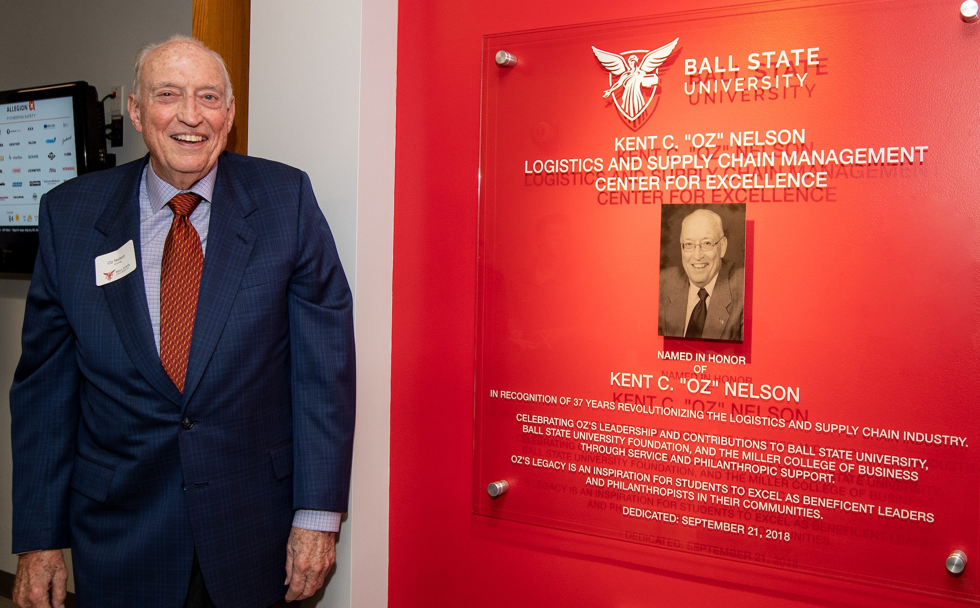 Oz Nelson wearing a blue suit standing next to the sign on the Kent C. "Oz" Nelson Logistics and Supply Chain Center for Excellence named in his honor. 