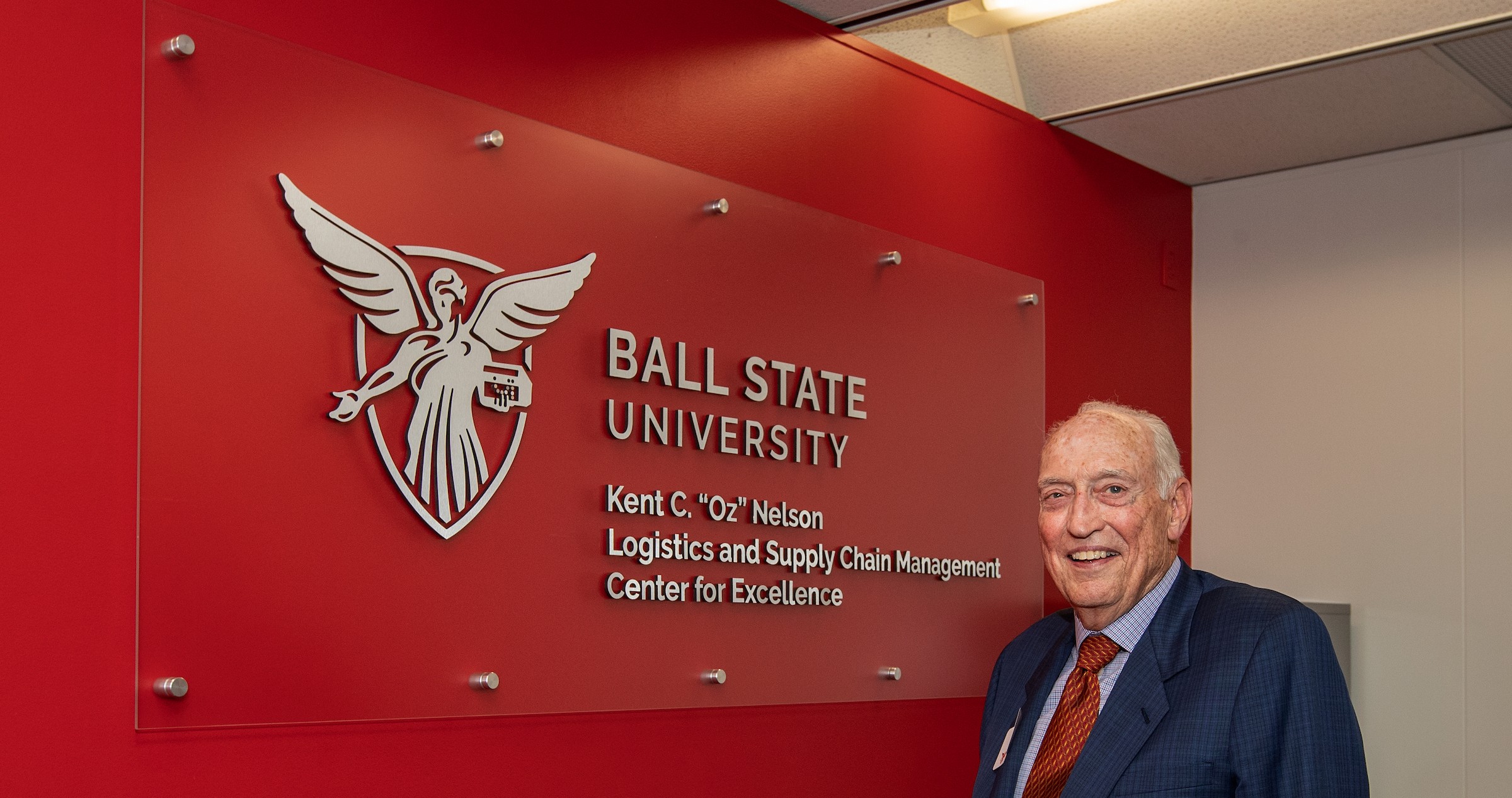  Gemini said An elderly man with white hair, identified as Kent "Oz" Nelson, stands smiling in front of a modern glass sign mounted on a bright red wall. He wears a blue suit jacket, a checkered shirt, and a red patterned tie.