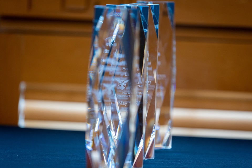 Several tall, clear glass or crystal awards with angular, faceted designs stand in a row on a dark blue tablecloth, with a softly blurred wood-paneled background behind them.