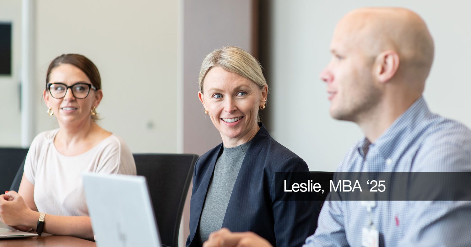 Three people sit at a table, two of them with laptops. A blonde woman looks at a man while he is talking. The text "Leslie, MBA, '25" in white on a gray box covers part of the right side of the image.