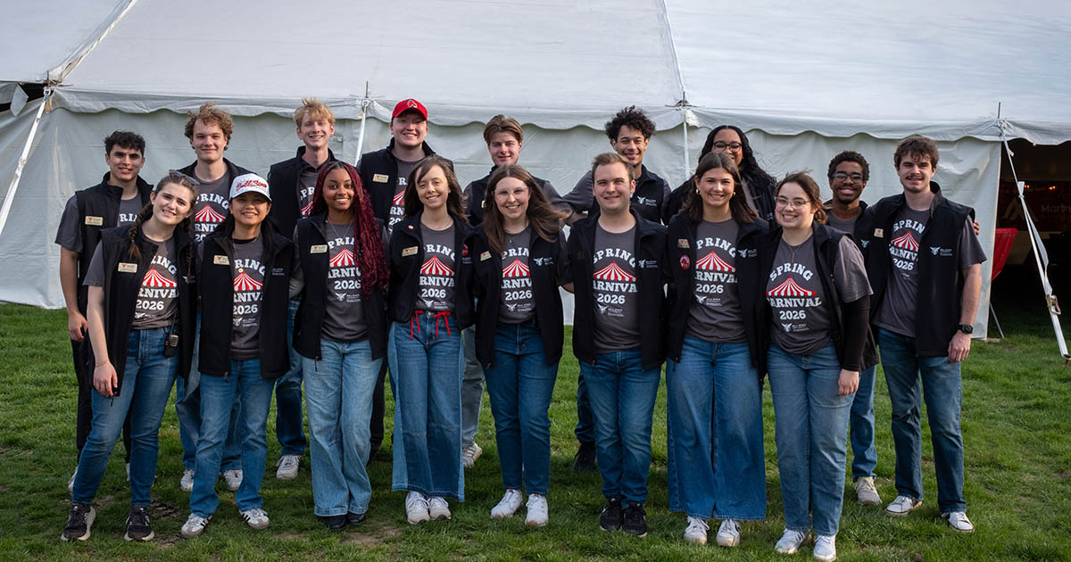 A groups of students, all wearing blue jeans, matching t-shirts, and vests with name tags, posing in front of a white tent for a group photo