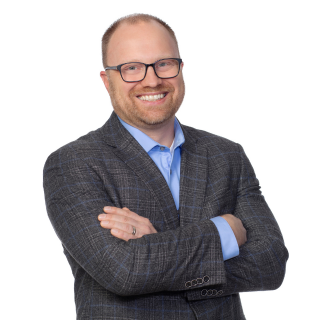 Mike Butler, wearing glasses and a grey suit and blue shirt while crossing his arms and smiling at the camera