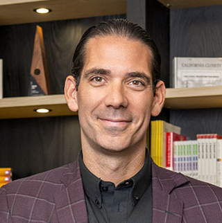 Head-and-shoulders portrait of Marcus Hall wearing a burgundy plaid blazer and black shirt, with bookshelves in the background. 