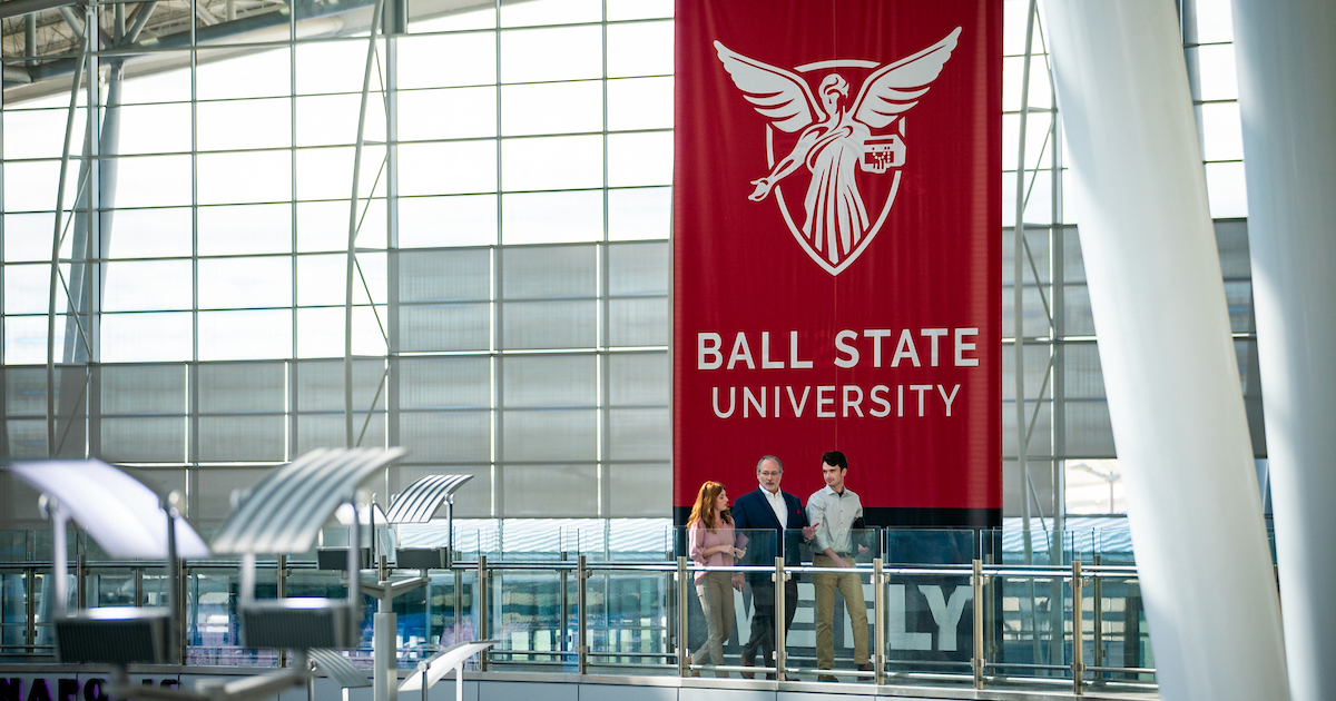 Three people walk in front of a large red banner with Ball State's logo that hangs from the ceiling in front of large windows at the Indianapolis International Airport 