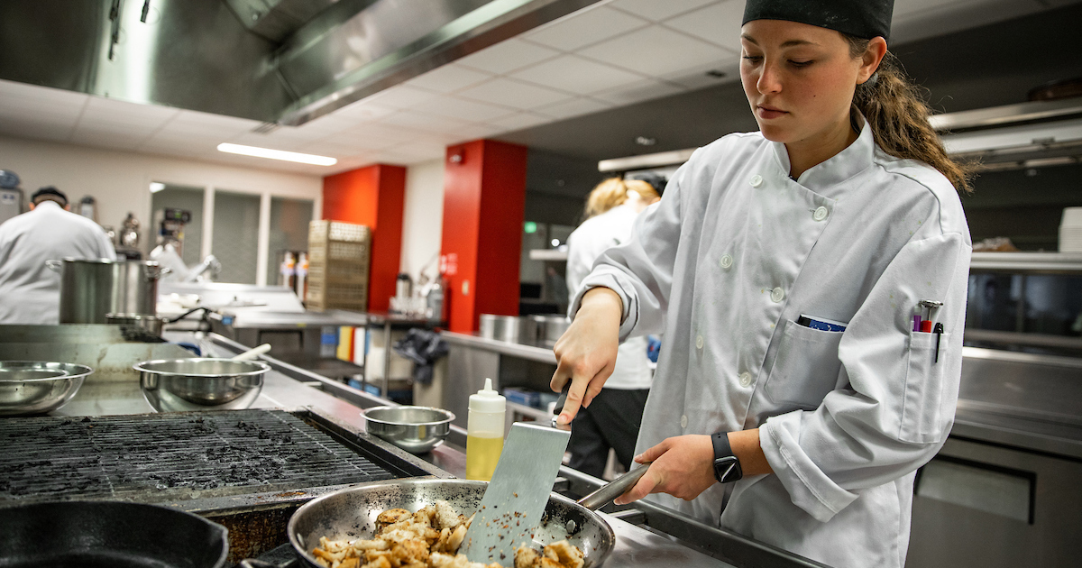 A young woman wearing a white cooking jacket and black cap stands in a busy kitchen while cooking food in a skillet