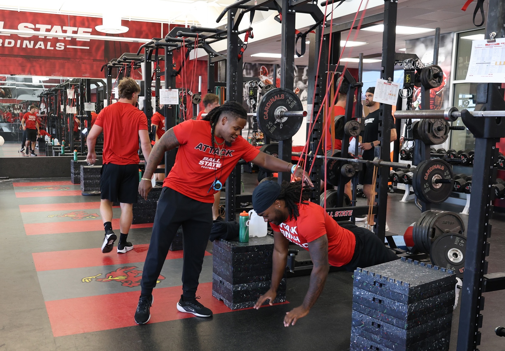 Kam Bryant leans over to coach a Ball State football player doing a push-up in the Cardinals weight room while teammates in red shirts walk behind them