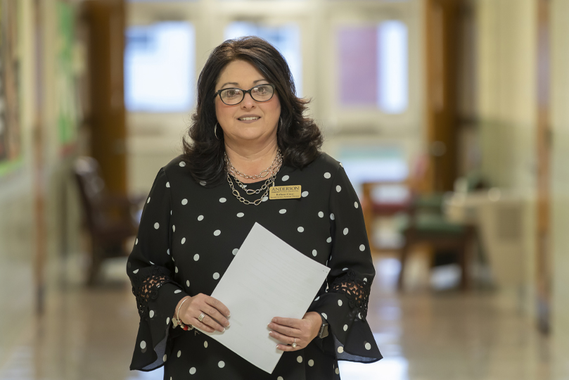 Woman with dark shoulder length hair, glasses standing in the hallway of a school with a black dress with white circles on it holding a piece of white paper with a gold name badge on her right pinned to her dress