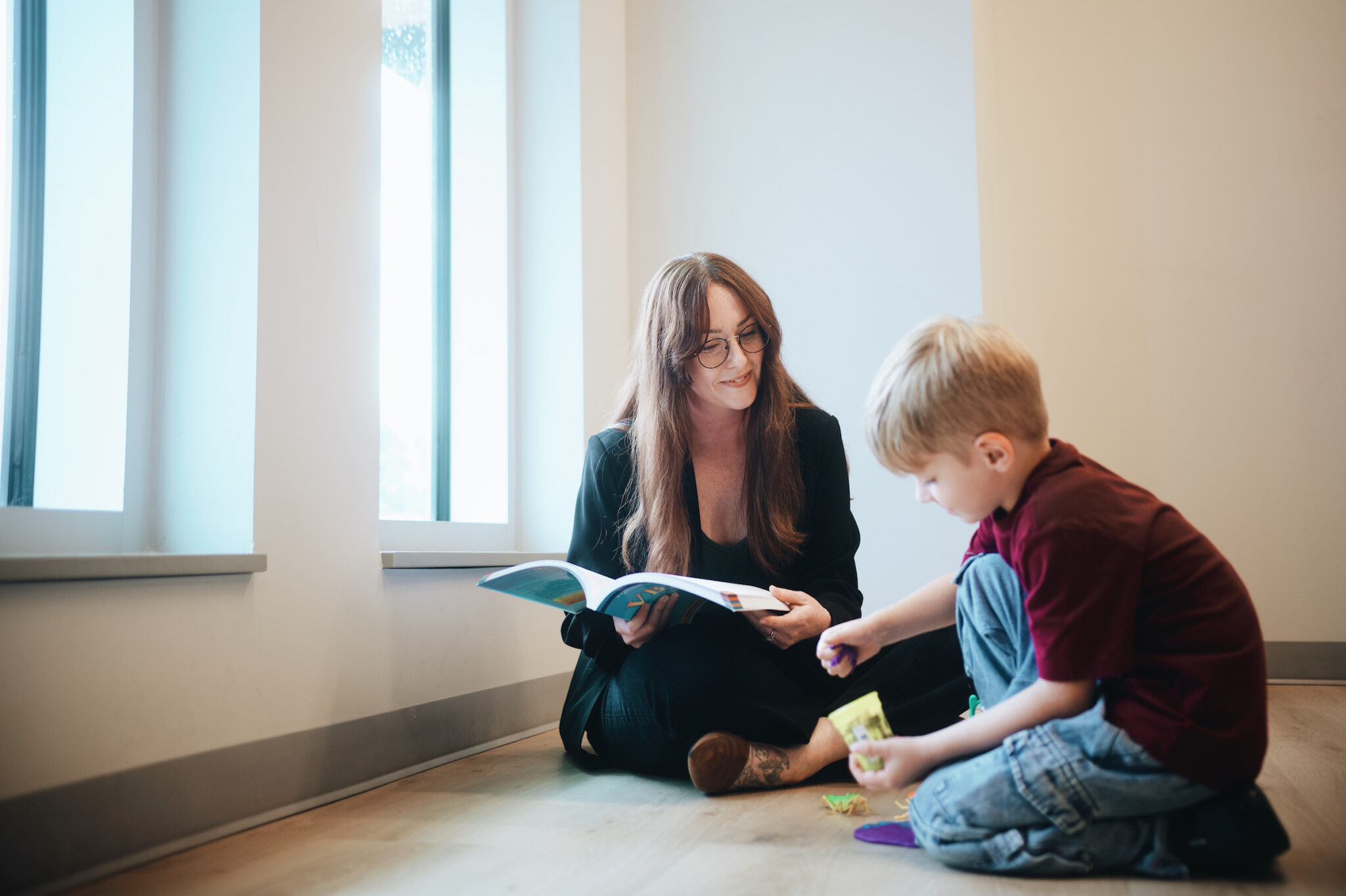 A woman sits on the floor next to a young boy by large windows. She holds an open book and smiles as the boy plays with small toys on the floor.