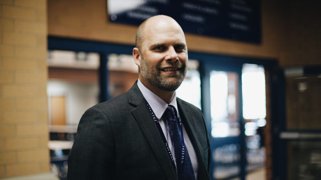 Adam Bontreger, man in a dark suit and tie with a lavender shirt standing in from of a classroom next to a brick column