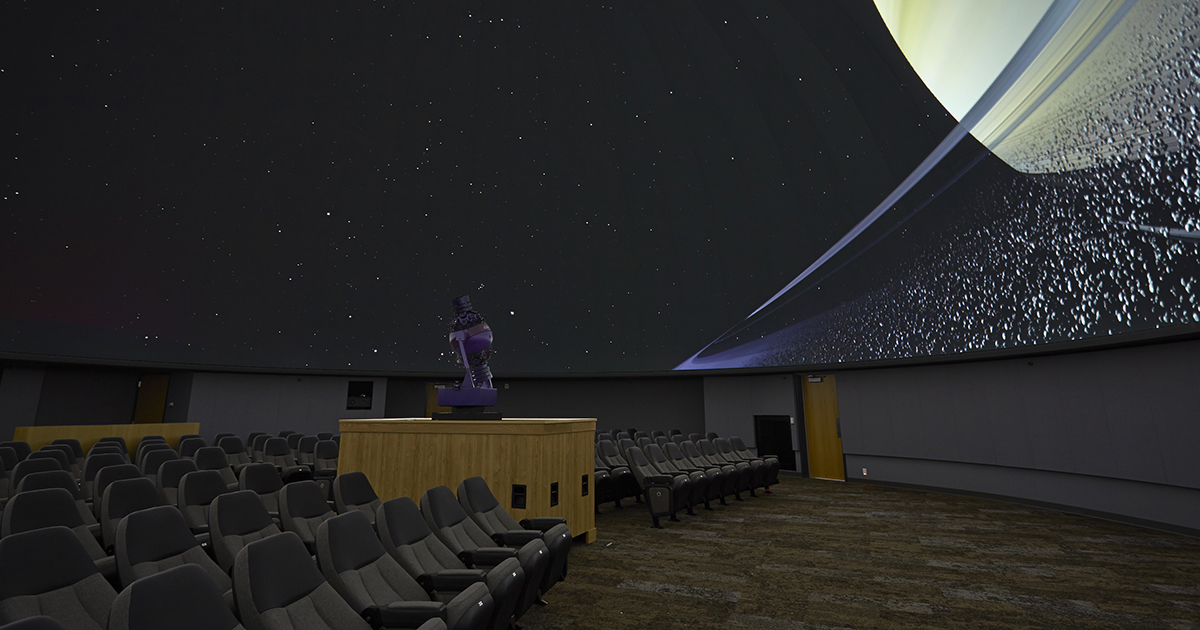 inside the planetarium, theater seats lined up with round star projector in the middle, Saturn surrounded by ring particles on dome., 