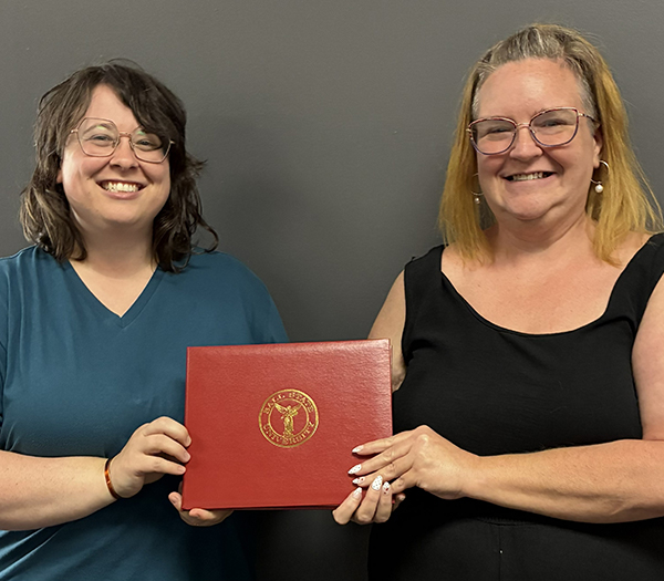 Two Ball State University employees smiling while holding a red Ball State presentation folder during a recognition photo.