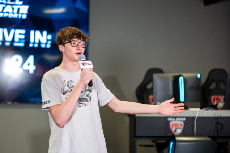 A student speaks into a microphone during an esports event, gesturing toward the audience while standing in front of gaming stations and a Ball State Esports display.