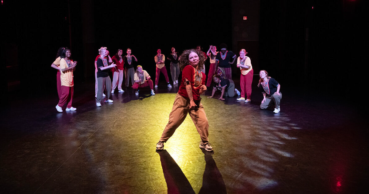A dancer performs confidently in the spotlight at center stage while fellow dancers surround and support the performance in a dark theater.