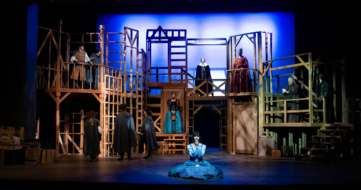 A large cast performs on an elaborate wooden scaffold set, with actors positioned on multiple levels under blue stage lighting.