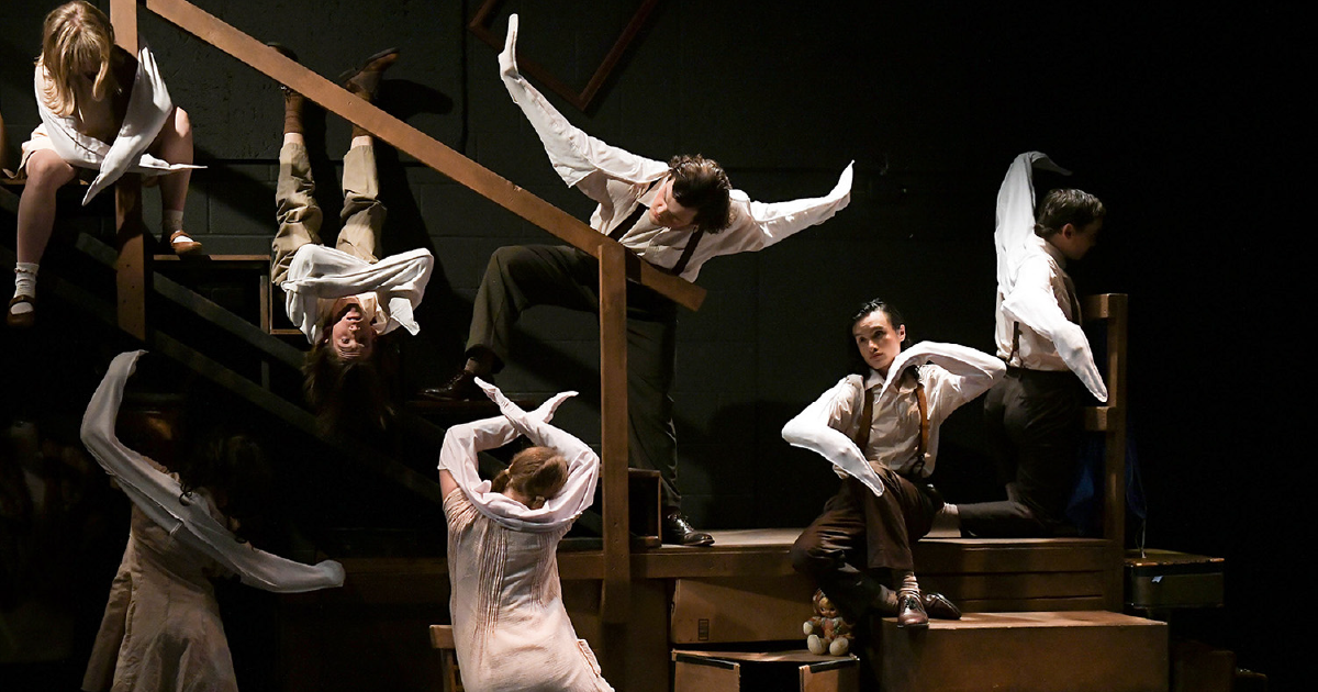 Actors in white costumes strike expressive, stylized poses across a multi-level staircase set during a dramatically lit stage performance.