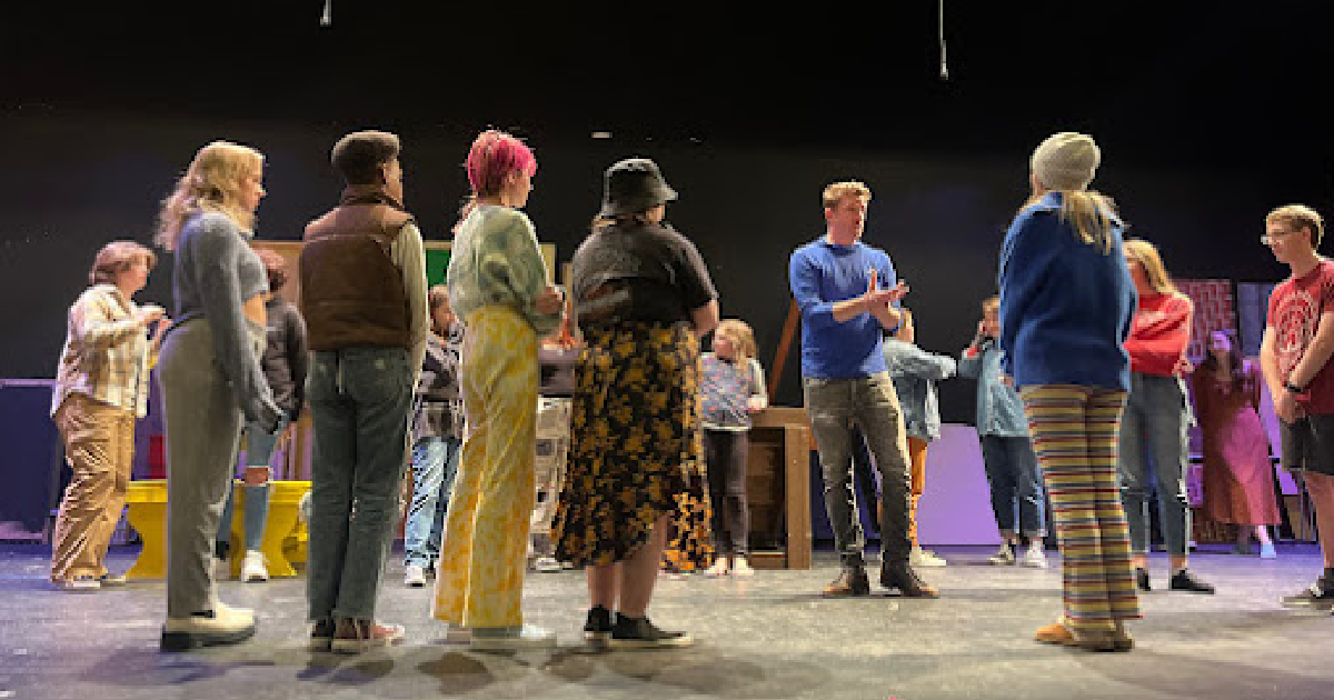 Several young adults stand on stage taking direction from one man in the center of the group.