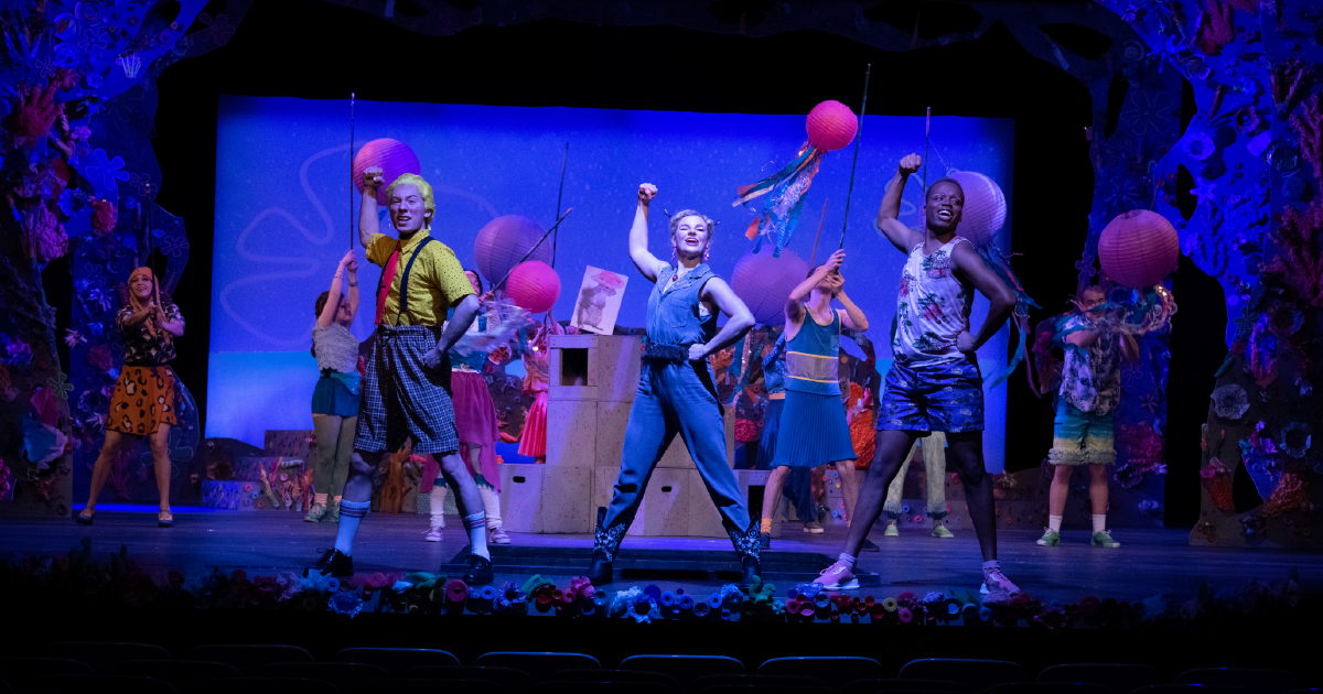 A colorful ensemble of performers sing and dance on a brightly lit stage decorated with whimsical props during a musical production.