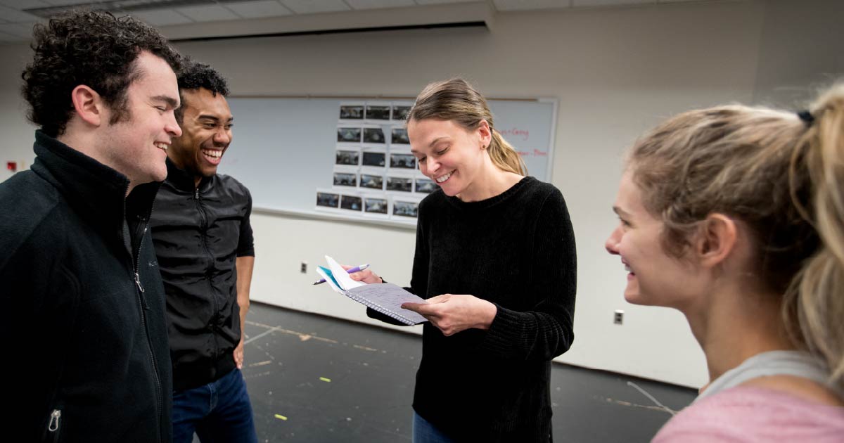 Three young adults stand in a classroom talking to Sutton Foster.