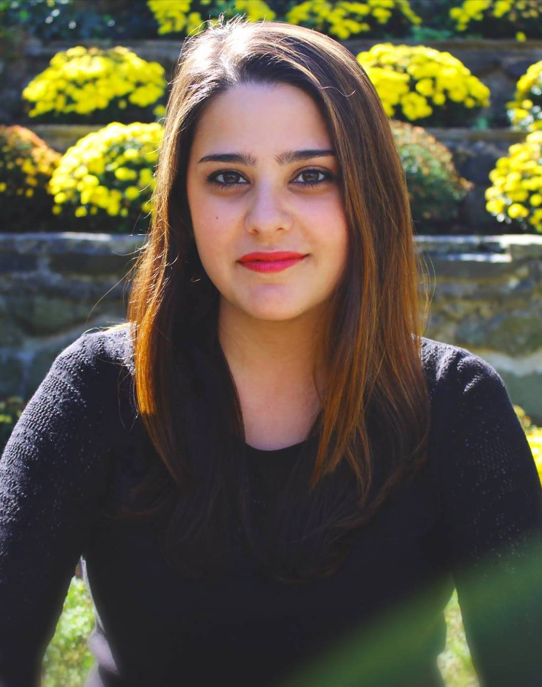 Catherine del Castillo Headshot - alumni theatre and dance - Latina and white biracial female with brown hair and red lipstick wearing a black longsleeved shirt. She is sitting in front of a pond with bushes of yellow flowers behind her.