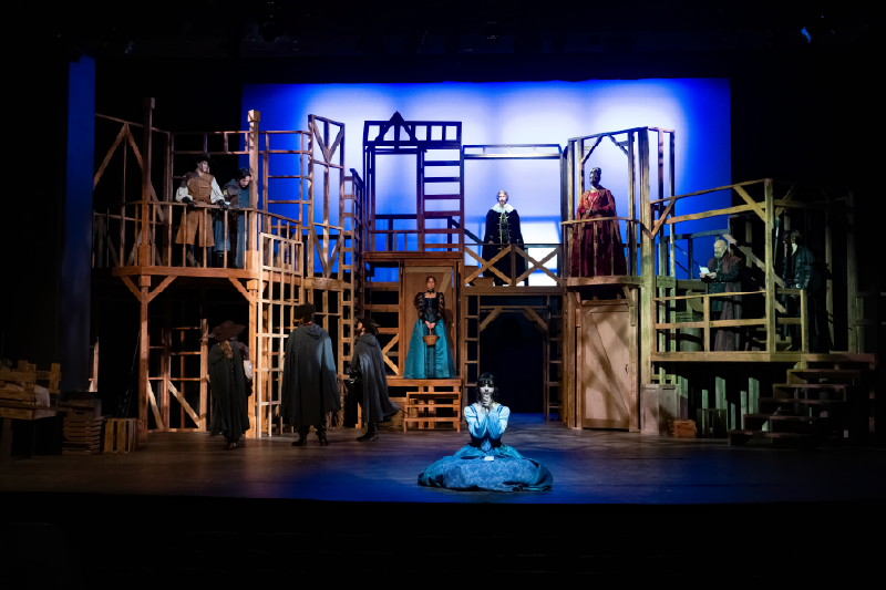 A large cast performs on an elaborate wooden scaffold set, with actors positioned on multiple levels under blue stage lighting.