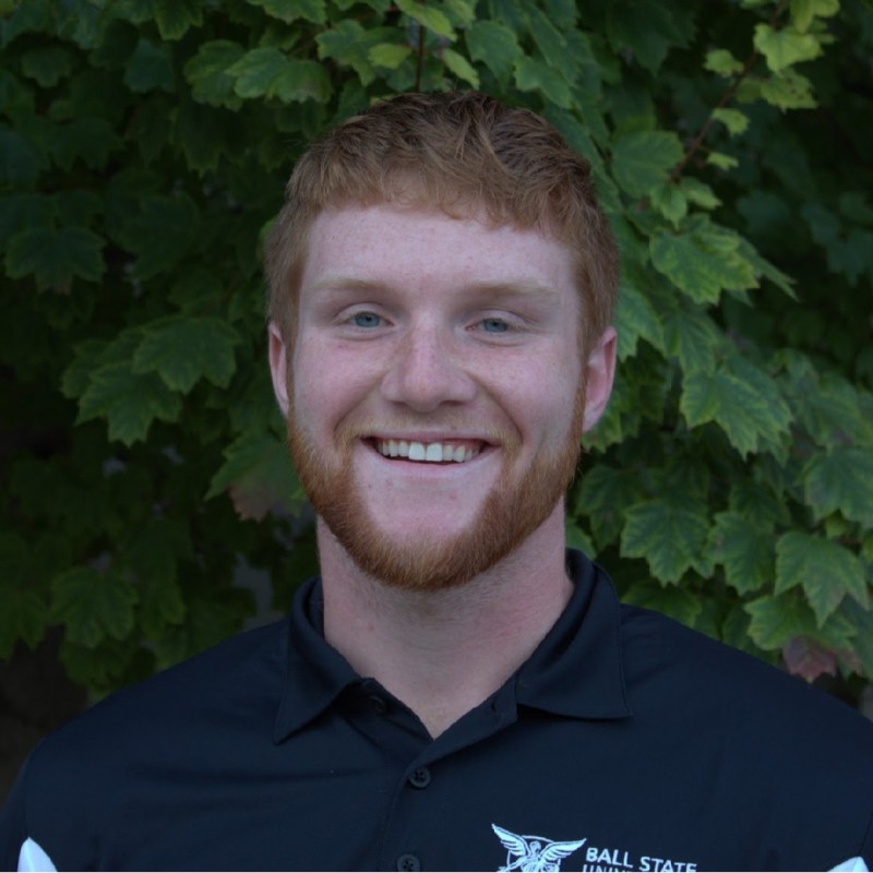 Cody Hauger wearing a black Ball State polo, smiling outdoors in front of green foliage.